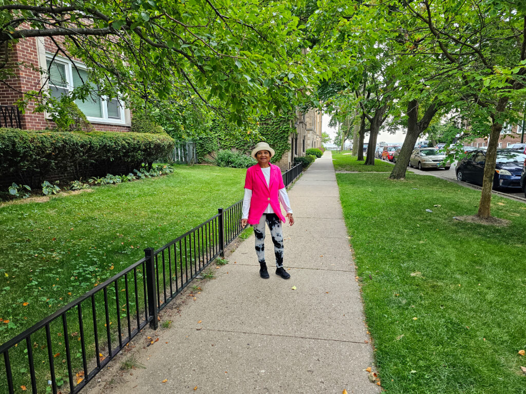 A woman in pink jacket standing on sidewalk next to fence.